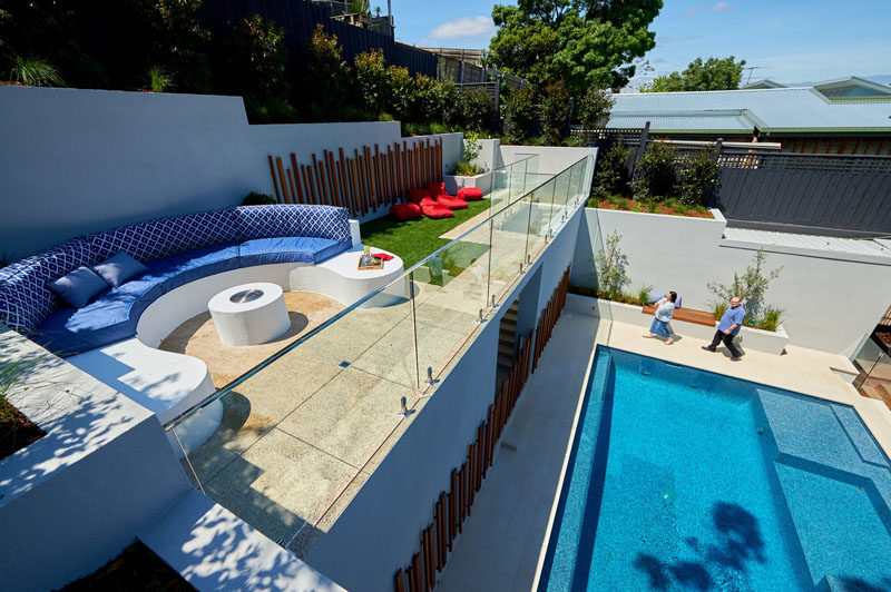 In this modern backyard, stairs lead from the pool level up to the lounge area with a built-in curved seating area, a touch of grass, and a glass railing that allows unobstructed views overlooking the pool and house. #ModernBackyard #LandscapeDesign #Garden #Pool #Landscaping