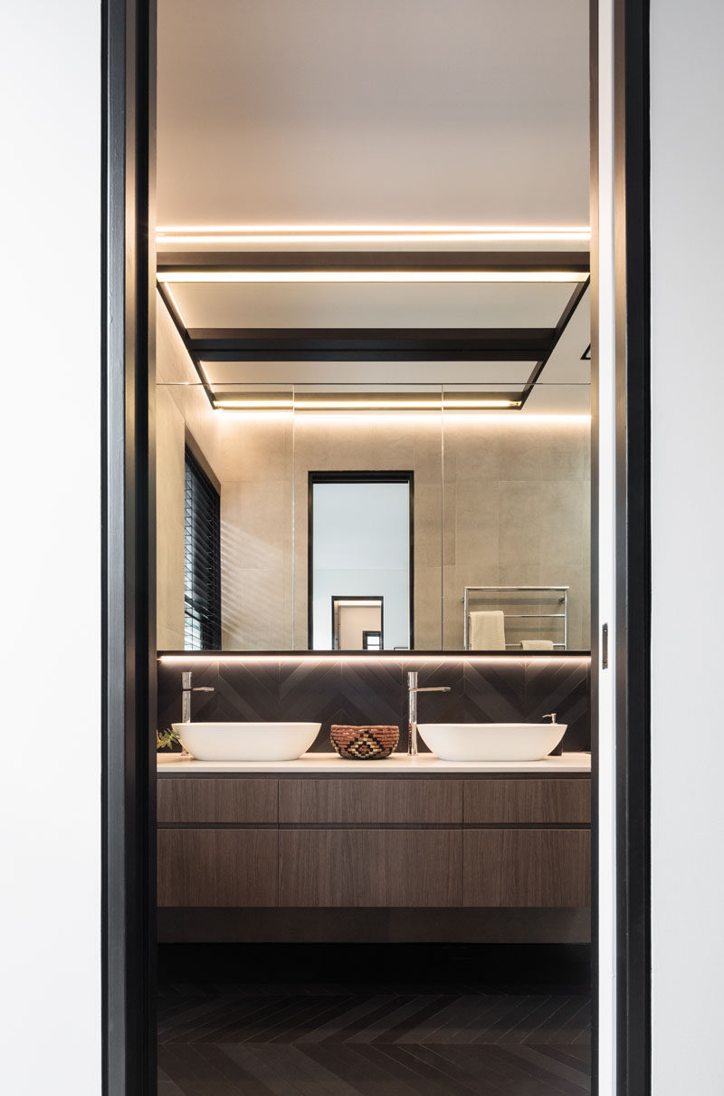 In this modern master ensuite bathroom, there's a dark chevron patterned backsplash and floor, while hidden lighting highlights the mirror. #EnsuiteBathroom #ChevronFloor #ModernBathroom #Lighting