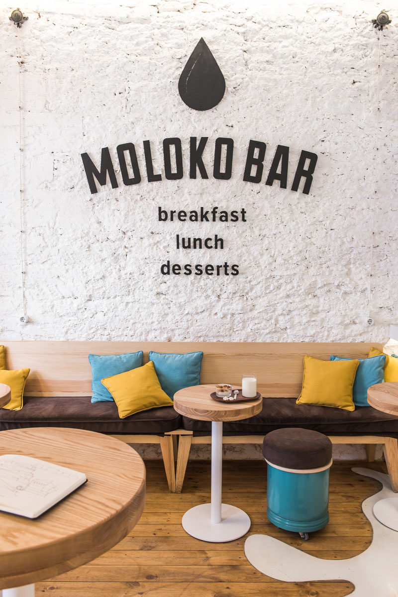 Inside this modern cafe, a textured wall that's been painted white, becomes a backdrop for the cafe's logo. Beneath the logo are long seats decorated with blue and yellow cushions. Little stools on wheels make it easy to create extra seating for small groups. #CafeDesign #CoffeeShop #InteriorDesign #Logo #GraphicDesign