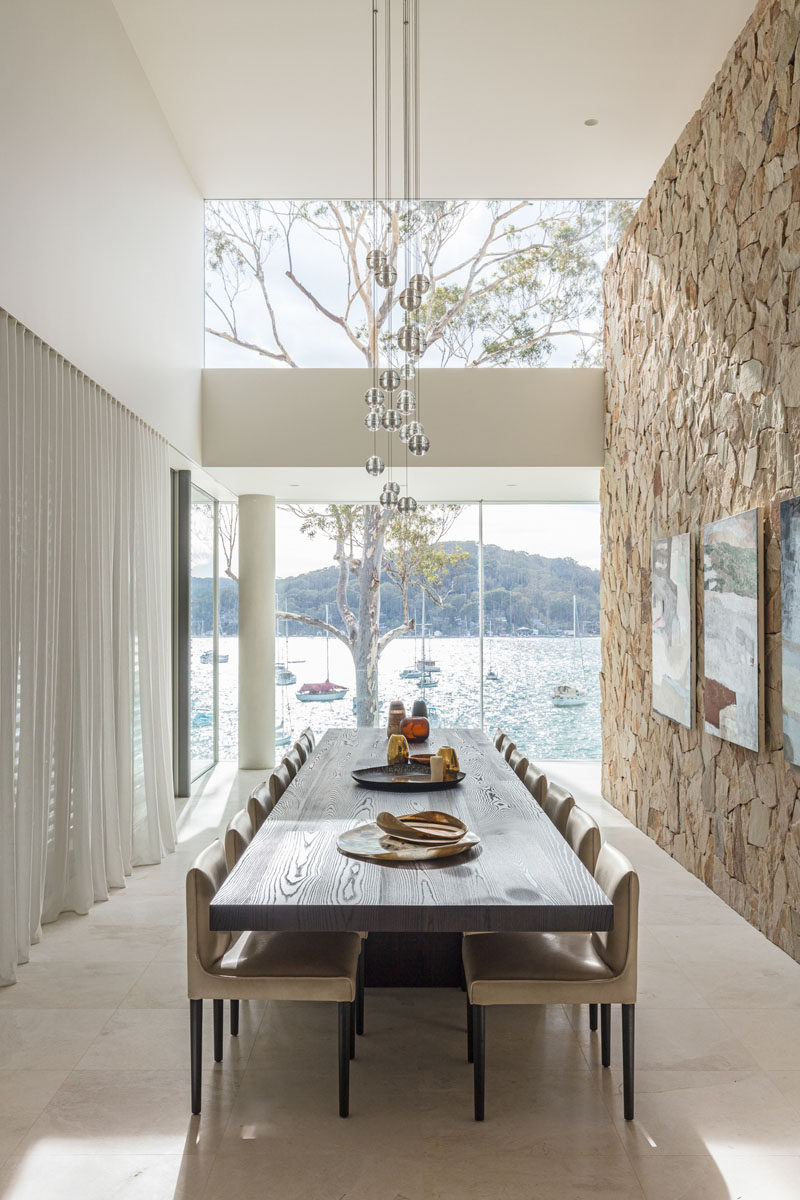 This formal dining room has a high ceiling, a sandstone accent wall, and a long 14-person dark wood dining table. #DiningRoom #SandstoneWall #InteriorDesign