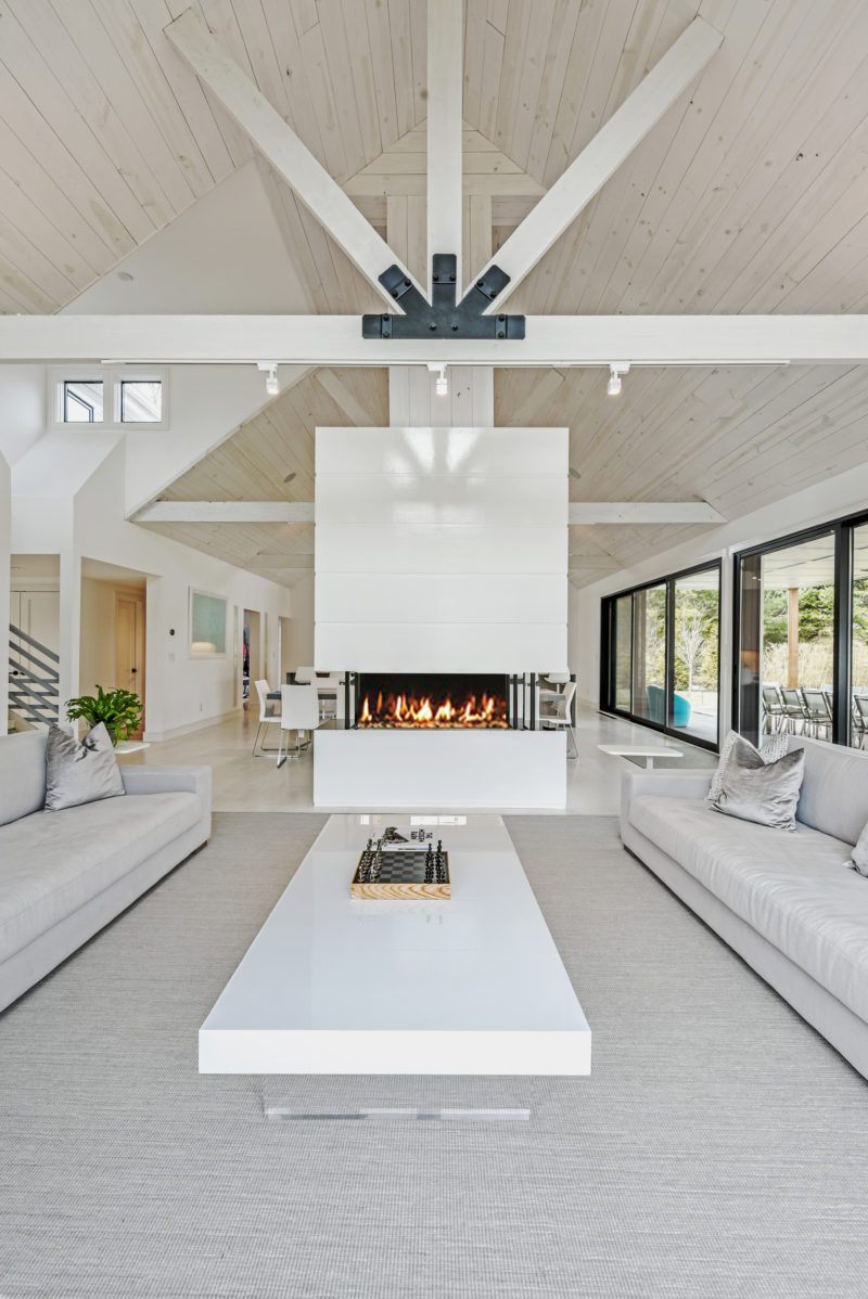 A double sided fireplace with a white surround draws the eye upwards to the exposed pitched ceiling and the structural supports of this modern barn interior. #Fireplace #ModernBarn #WhiteInterior