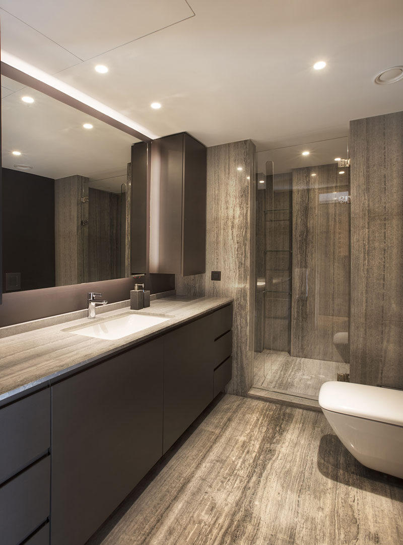 In this modern bathroom, stone covers the flooring and the shower, as well as the vanity, which has plenty of counter space and a backlit mirror. #ModernBathroom #BathroomDesign