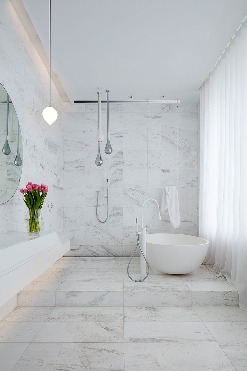 In this modern white bathroom, the walls and floor are covered in tiles, with the freestanding bathtub positioned on a raised platform. #ModernBathroom #BathroomDesign #WhiteBathroom