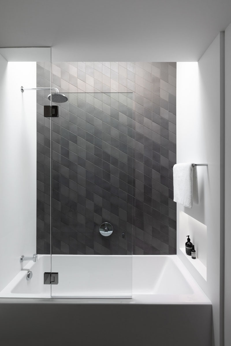 In this bathroom, dark tile has been used to create an accent wall, and a small built-in shelf with lighting at the end of the bath provides a place for storing bottles. #Bathroom #ModernBathroom #BathroomDesign