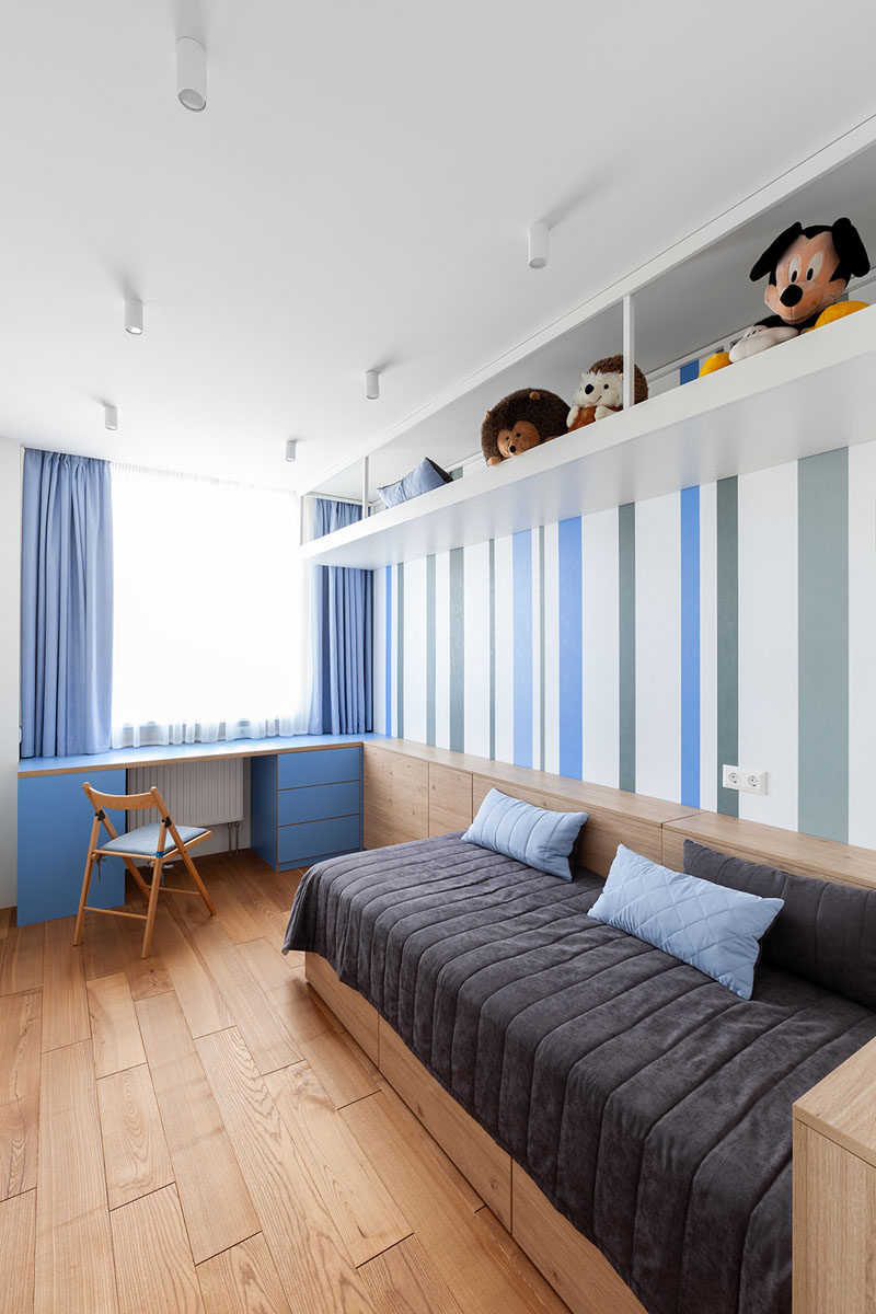 In this modern bedroom, a blue desk has been positioned in front of the window, while the bed doubles as a couch, and a a long shelf runs along the ceiling. #BedroomDesign #ModernBedroom