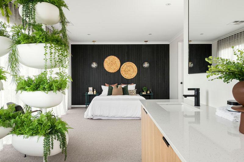 In this modern master bedroom, a black wood accent wall draws the eye to the bed, that looks towards an open ensuite bathroom with a decorative plant stand providing some privacy for the freestanding bathtub. #MasterBedroom #BlackAccentWall #InteriorDesign