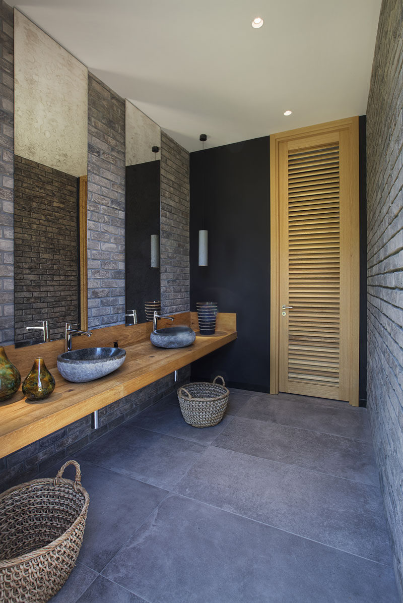 This bathroom has brick walls, a long wood vanity, and tall mirrors that meet the ceiling. #BathroomDesign #ModernBathroom