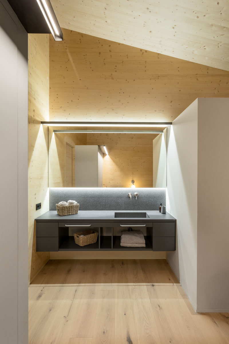 In this modern bathroom, grey cabinetry stands out against the wood walls and ceiling, while a backlit mirror and minimalist lighting highlight the vanity. #ModernBathroom #Lighting #BathroomDesign