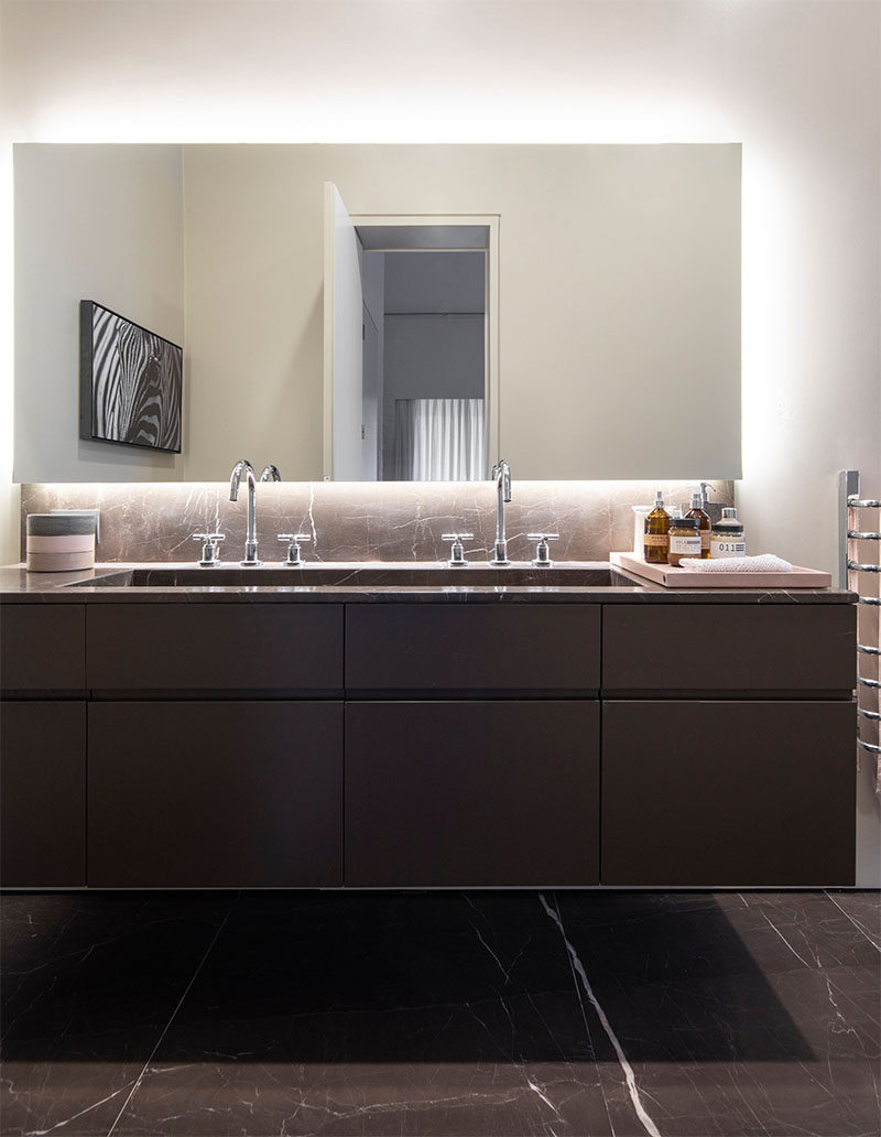 In this modern bathroom, a trough sink vanity with two faucets sits below a backlit mirror. #ModernBathroom #BathroomDesign #BacklitMirror