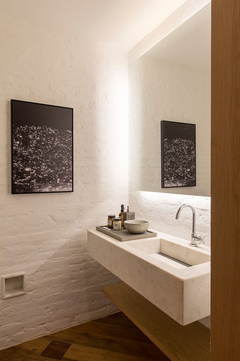 In this modern bathroom, a light-colored stone vanity with a built-in sink floats above a wood shelf, while a backlit mirror is mounted above. #ModernBathroom #BacklitMirror