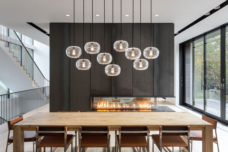 In this dining area beside a black steel fireplace, a wood dining table is positioned under a collection of pendant lights hung at different heights. #ModernDiningRoom #Fireplace