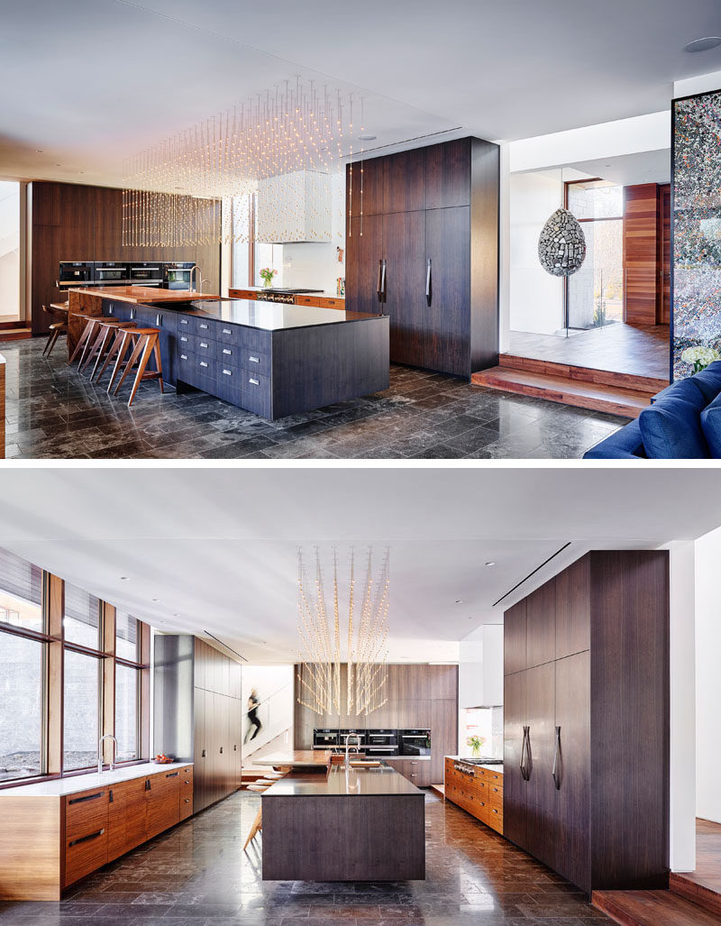 In this modern kitchen, dark wood cabinetry has been paired with lighter wood accents, while a long kitchen island is positioned centrally in the space. Above the island is a decorative light installation that brightens the kitchen. #ModernKitchen #KitchenDesign