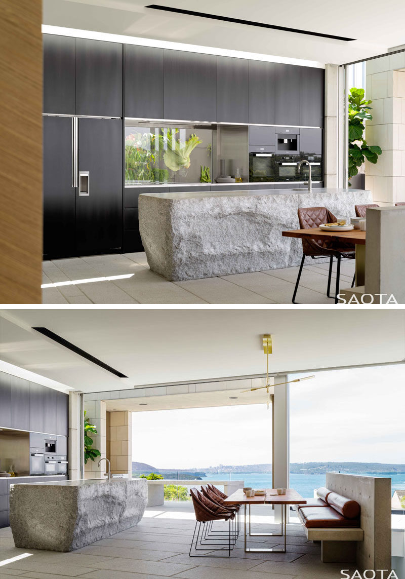 In this modern kitchen, a hand-cut grey granite kitchen island makes a statement, while dark minimalist cabinetry and glass backsplash line the wall. Also included in the same room as the kitchen, is a secondary, more casual, dining area. #ModernKitchen #KitchenDesign #Dining