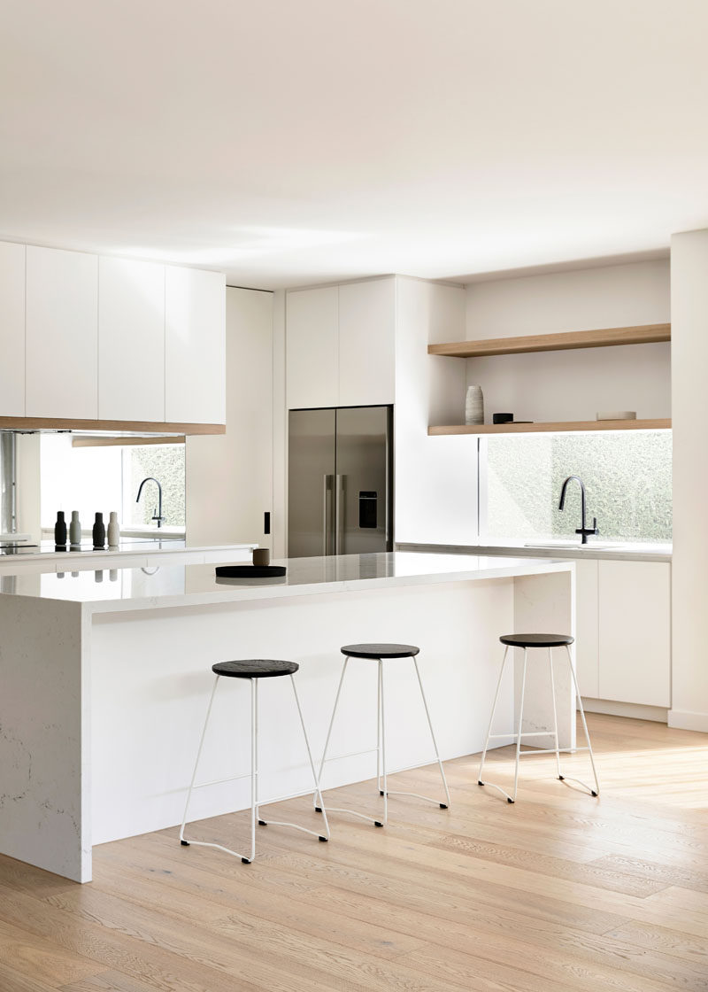 This modern kitchen has bright white cabinets with light wood detailing. A window looking out to the side garden acts as a backsplash, and a mirror below the other cabinets helps to reflect light throughout the space. #WhiteKitchen #WindowBacksplash #MirroredBacksplash