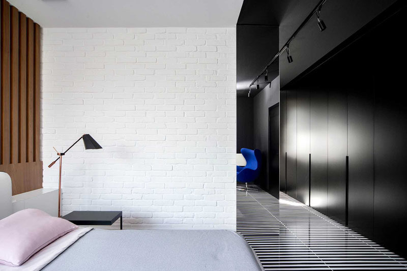 Widawscy Studio Architektury have designed a modern master bedroom that features a white brick wall, a wood slat accent wall, black and white striped flooring, black closets, and pop of bright blue in the form of an chair. #MasterBedroom #StripedFlooring #BlackClosets