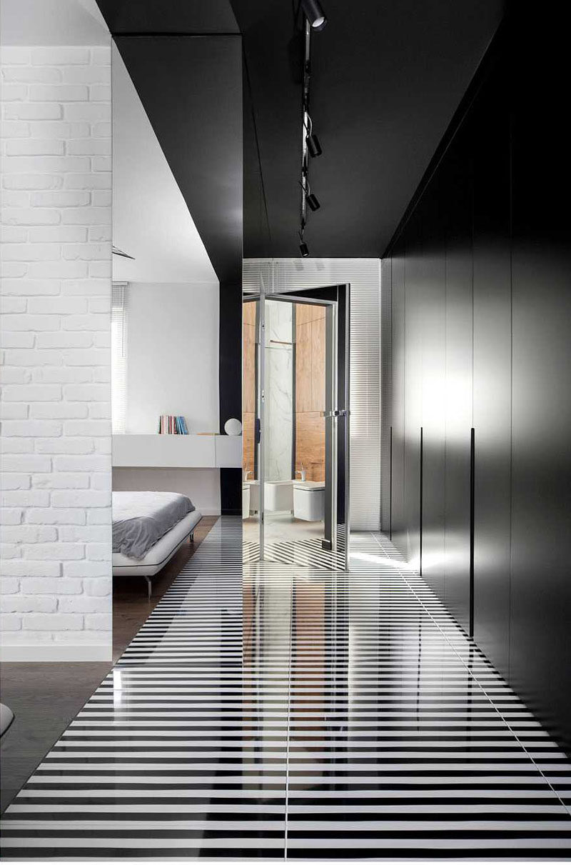 Widawscy Studio Architektury have designed a modern master bedroom that features a white brick wall, a wood slat accent wall, black and white striped flooring, black closets, and pop of bright blue in the form of an chair. #MasterBedroom #StripedFlooring #BlackClosets