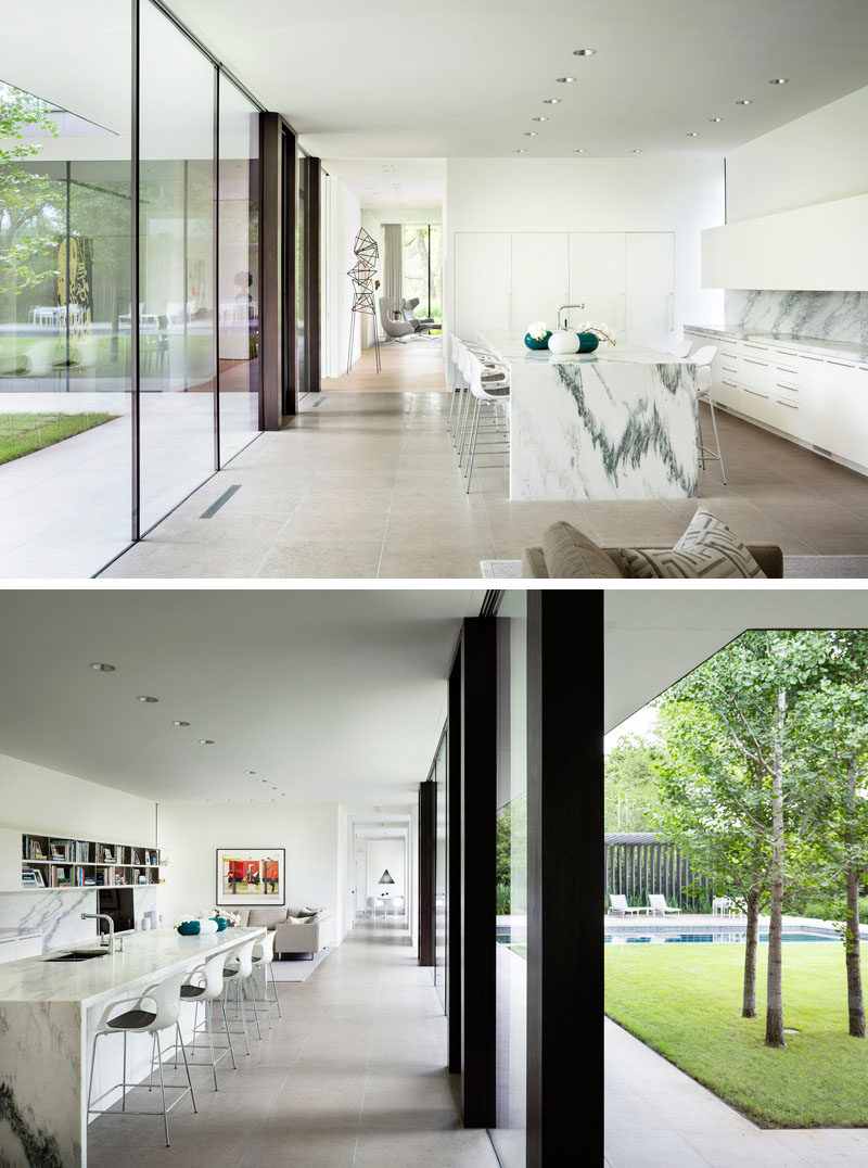 This modern kitchen has a large island, minimalist white cabinets, and floor-to-ceiling windows. Beside the kitchen is a casual den/living room. #WhiteKitchen #ModernKitchen #KitchenDesign