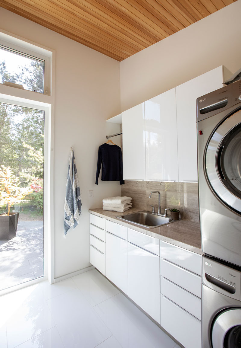 This modern laundry room, with its high ceilings, has plenty of storage in glossy white cabinets, a place to hang clothes and stackable washer/dryer. #ModernLaundry #LaundryRoom