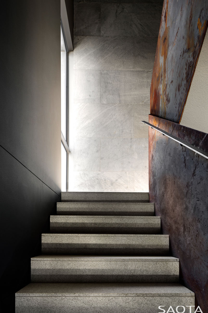 The staircase in this modern house consists of light grey granite stair tiles with crude steel side balustrade sheets that have been left to rust naturally, while the walls are matte silver grey granite. #ModernStairs #SteelBalustrade