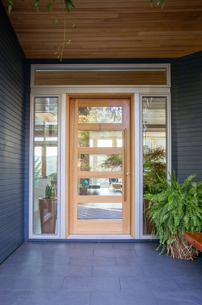 A wood and glass front door surrounded by windows welcomes visitors to the home. #FrontDoor #ModernFrontDoor