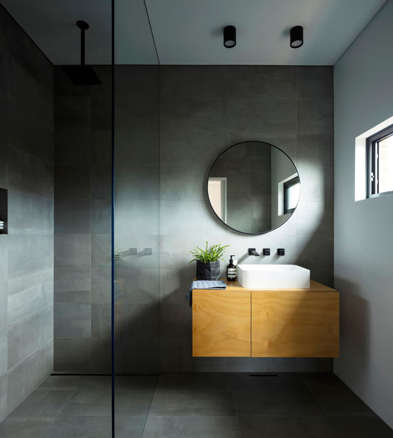 This modern master ensuite bathroom has dark grey tiled walls and floor, a light wood vanity, and a round mirror. #MasterBathroom #BathroomDesign #GreyTiles