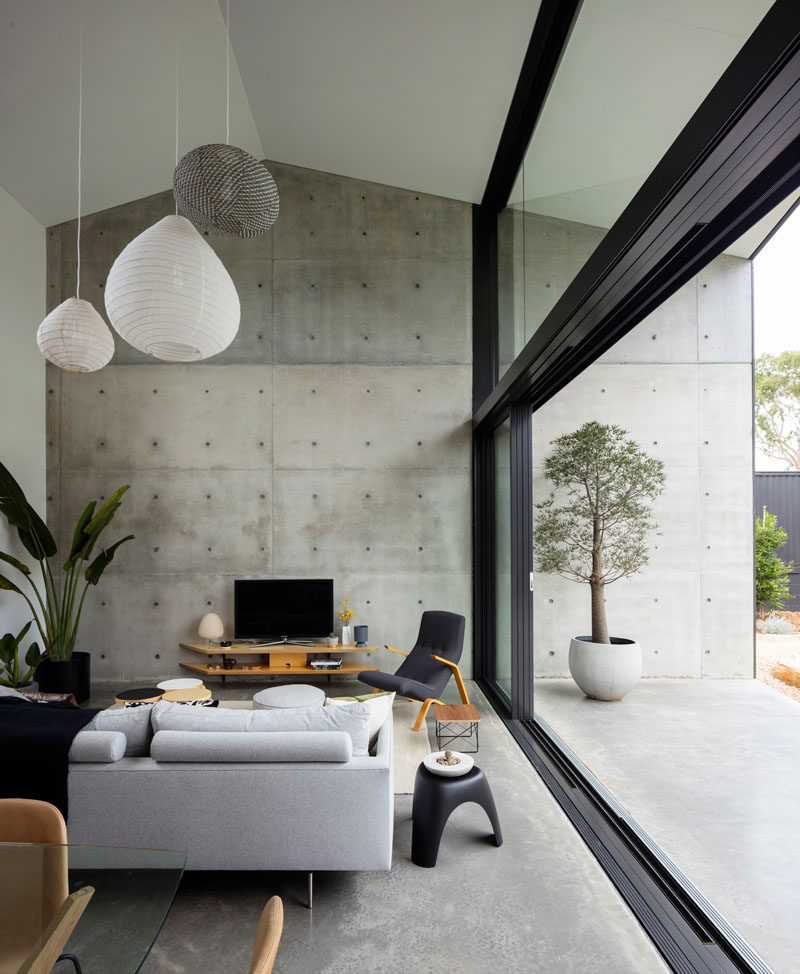 In this modern living room area, a double-height ceiling creates an open feeling, while a large exposed concrete wall continues from the interior to the exterior of the home. #LivingRoom #ConcreteWall