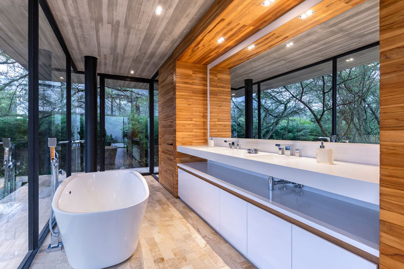 In this modern master bathroom there's a freestanding bathtub, and a long vanity with a large mirror that reflects the treetops. #MasterBathroom #BathroomDesign #ModernBathroom