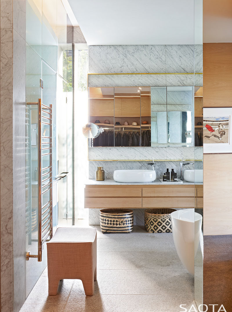 This modern bathroom features features gold accents, stone, glass, wood and a white sinks that complement the white freestanding bathtub. #ModernBathroom #BathroomDesign