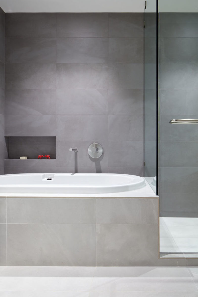 In this modern bathroom, the bathtub has been built-in and large grey tiles that contrast the white bathtub, cover the walls and bathtub surround. #GreyBathroom #ModernBathroom #BuiltInBathtub