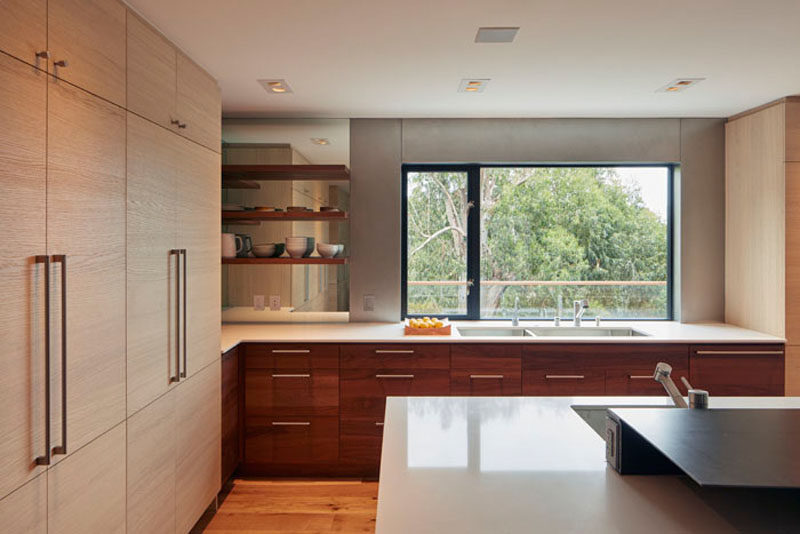 In this modern kitchen, dark and light wood cabinetry are featured, while a window frames a view of the trees, and a mirror reflects the floating shelves and light, making the corner brighter.