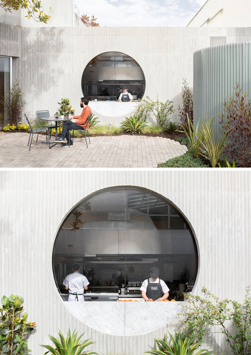 In this restaurant's courtyard, there's a stone patio with seating, that's surrounded by curved landscaping, while a round window provides a glimpse into the kitchen. #Courtyard #Window #Restaurant