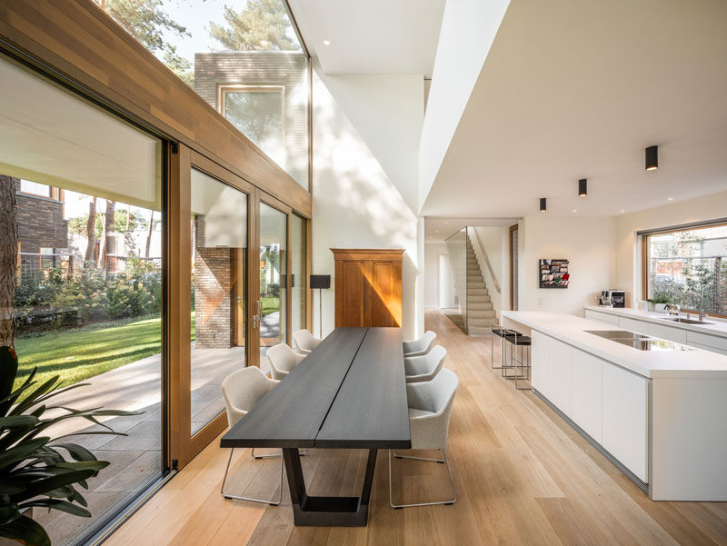 This modern house has a double-height ceiling above dining table, large windows for plenty of natural light, and wood framed sliding doors to connect to the outdoors. #DiningRoom #WhiteKitchen #DoubleHeightCeiling #Windows #SlidingDoors