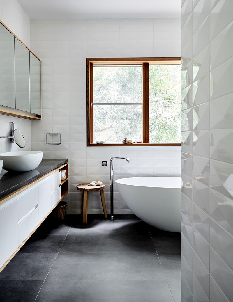 In this modern bathroom, the walls have been covered in a three-dimensional white tile, creating visual interest and contrasting the grey tiles and vanity counter. #BathroomDesign #ModernBathroom