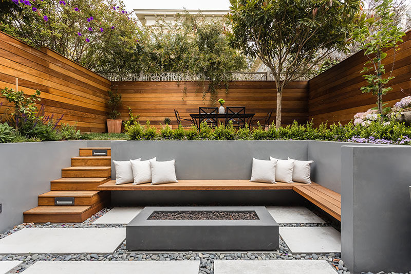 On the lower level of this modern backyard, there's custom-colored concrete walls with a built-in wood bench that fits into the corner and sits beside the firepit. On the ground, pavers are surrounded by riverstone, while wood stairs lead to the upper level with an area for alfresco dining. #ModernLandscaping #ModernBackyard #MultiLevelBackyard