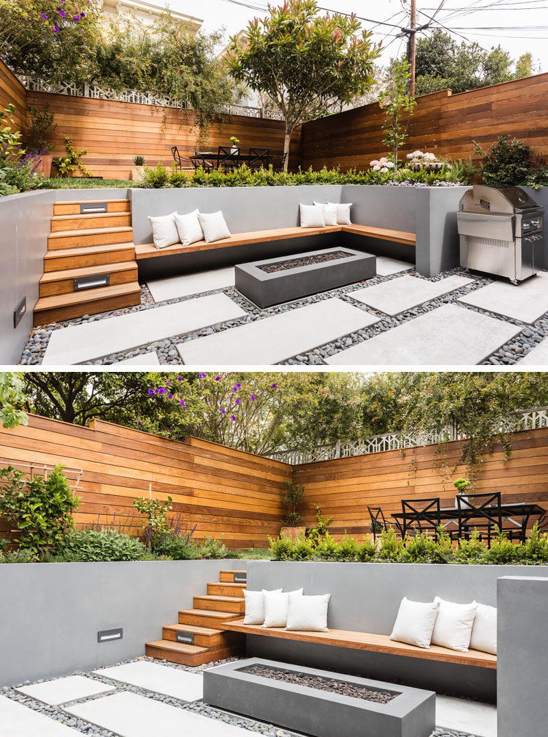 On the lower level of this modern backyard, there's custom-colored concrete walls with a built-in wood bench that fits into the corner and sits beside the firepit. On the ground, pavers are surrounded by riverstone, while wood stairs lead to the upper level with an area for alfresco dining. #ModernLandscaping #ModernBackyard #MultiLevelBackyard