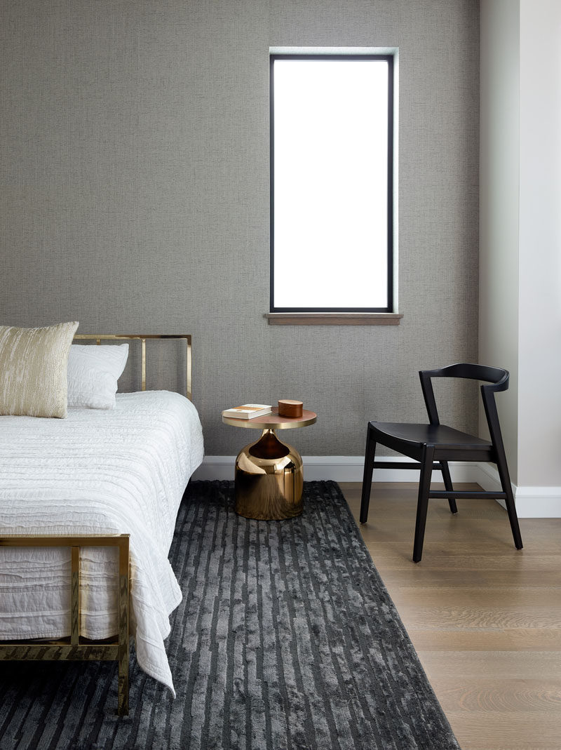 In this modern bedroom, a grey rug complements the grey accent wall, while the black chair ties in with the window frame, and the gold accents add a metallic touch. #ModernBedroom #BedroomDesign