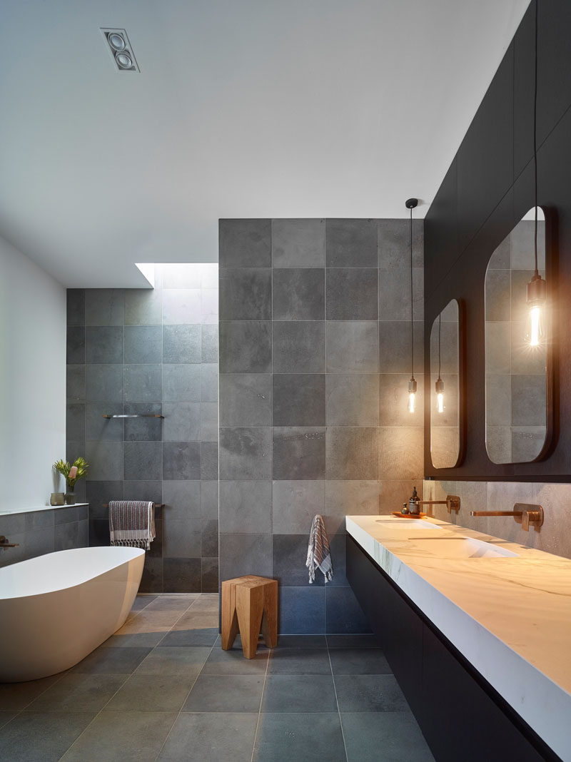 This modern bathroom features a black, white and grey color palette, with a walk-in shower, a freestanding bathtub, and a large dual-sink vanity that sits below two mirrors with pendant lights either side. #BathroomDesign #ModermBathroom