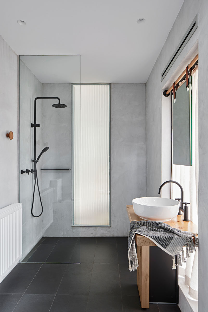 In this modern bathroom, a mirror in front of the window hangs from straps attached to a wood rod, while a floor-to-ceiling glass shower screen separates the shower from the rest of the bathroom. #ModernBathroom #BathroomDesign