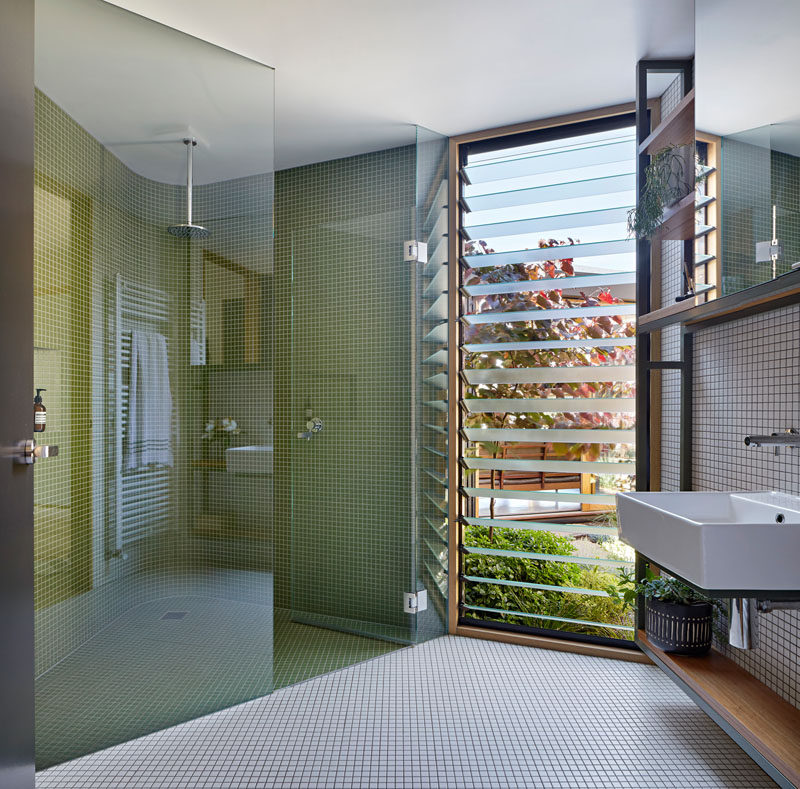 This modern bathroom has louvre windows, and a glass enclosed shower with a curved wall that's covered in small green tiles. #BathroomDesign #GreenTiles #ModernBathroom