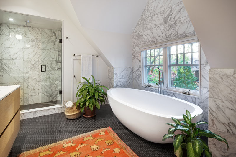 In this contemporary bathroom, high ceilings create a lofty feeling, while the freestanding white bathtub is positioned in front of the window that has tree views. #BathroomDesign #ModernBathroom