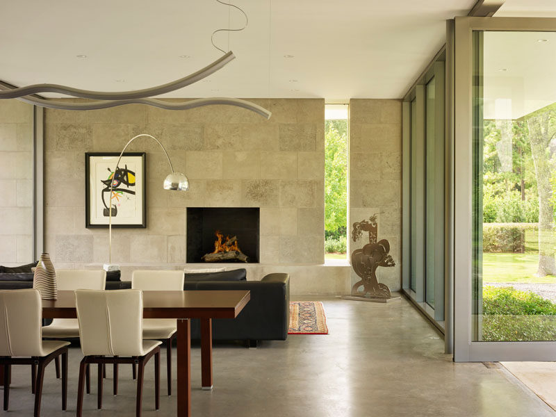 Inside this modern house, a limestone wall surrounds a fireplace, while the floor-to-ceiling windows provide views of garden. #LimestoneWall #InteriorDeisgn