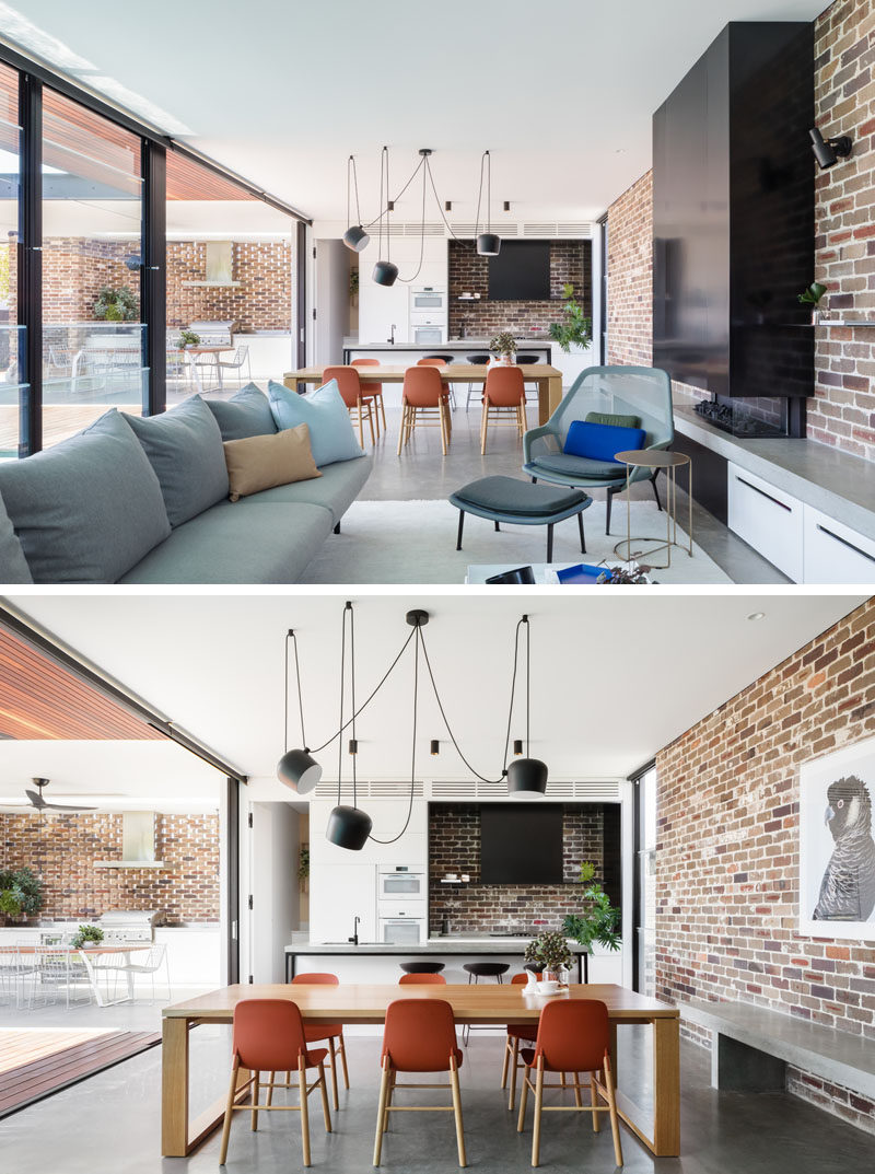 In this open plan living room, dining room, and kitchen, there's a recycled brick wall that wraps around the room, and provides a backdrop for the black fireplace and concrete bench. #InteriorDesign #Brick #OpenPlanInterior