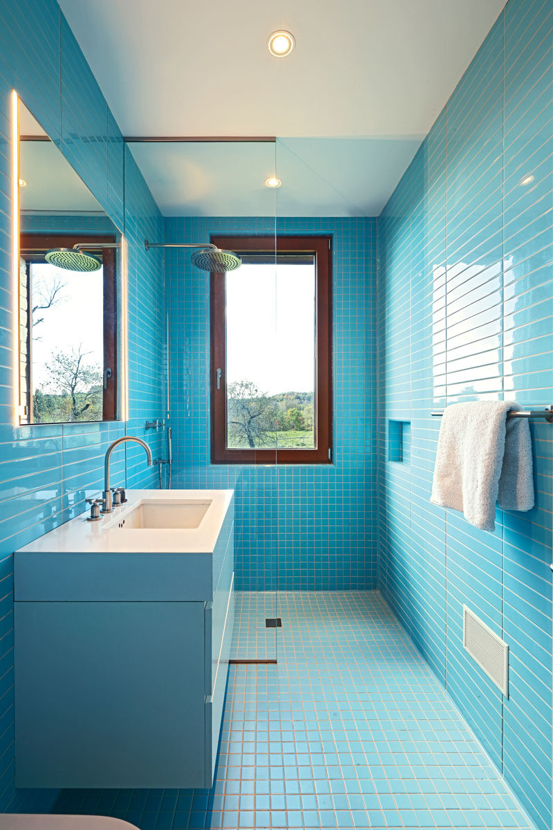 In this modern bathroom, bright blue rectangular and square tiles cover the walls and floor, contrasting the wood window frame and white vanity. #BlueBathroom #ModernBathroom #BathroomDesign