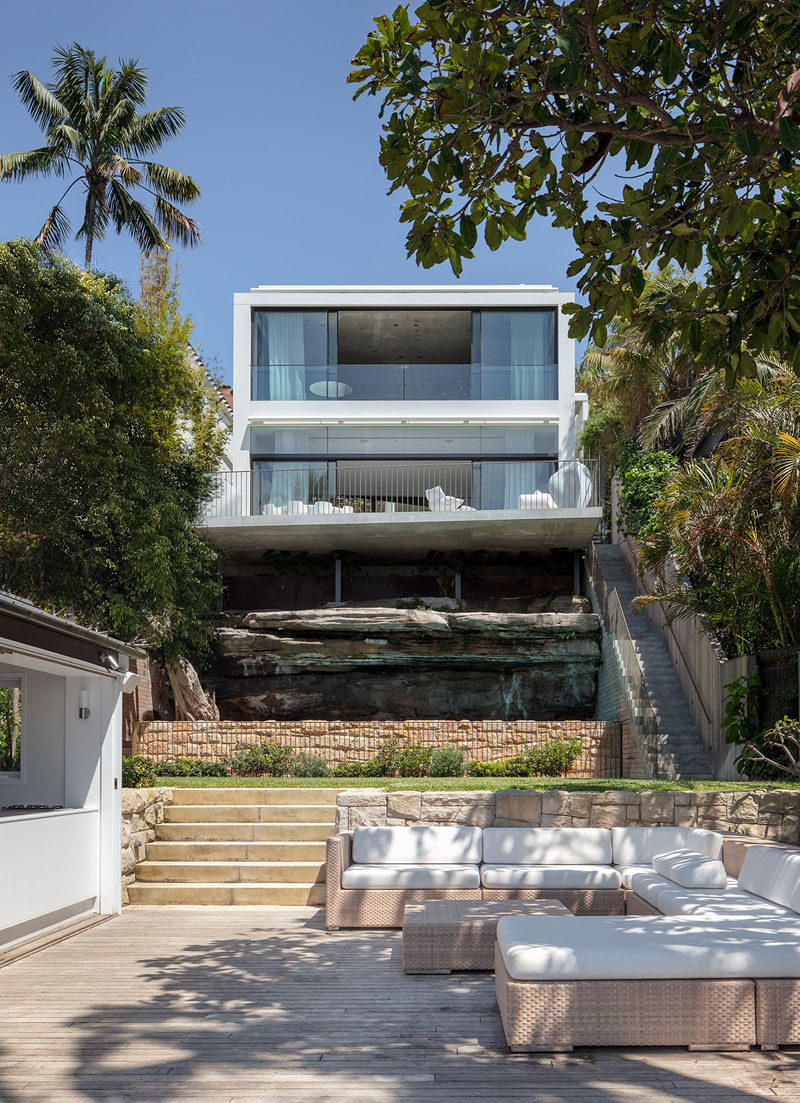 As this modern house sits on a sloped lot, there are stairs located on the side of the house, that connect to a tiered backyard with an outdoor lounge area close to the harbor. #ModernHouse #Landscaping #OutdoorLounge