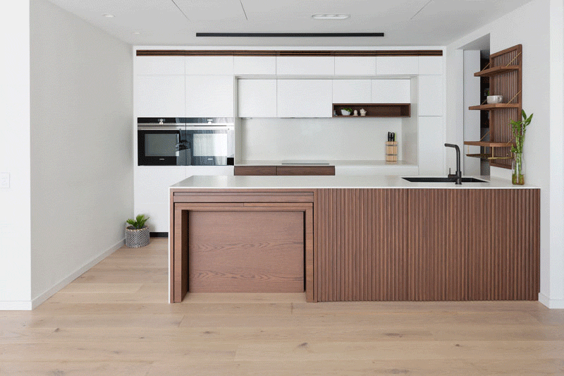 This modern kitchen features custom stained American Oak, corian countertops, and pull-out dining table, which sits flush within the kitchen bench and extends into a large dining table. #ModernKitchen #HiddenDiningTable #BuiltInDiningTable