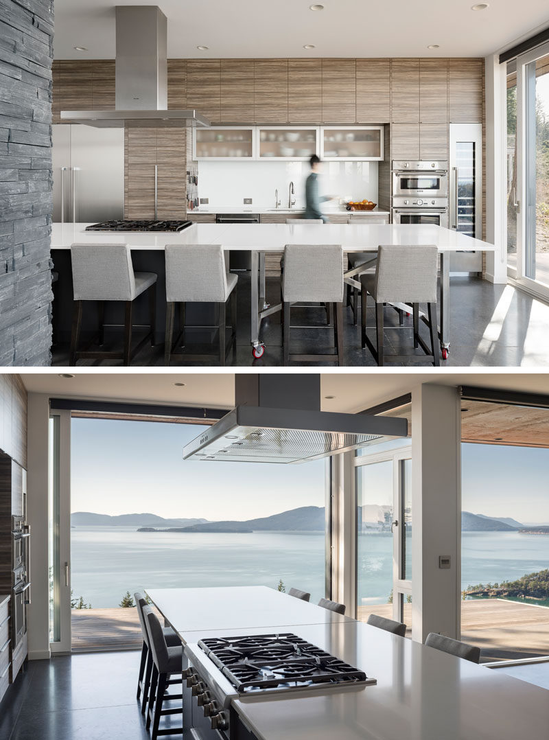 In this modern kitchen, minimalist hardware-free wood cabinets line the wall, and a large white kitchen island provides plenty of counterspace. A movable dining table has been designed to extend the kitchen island, or be wheeled outside onto the deck to be used for alfresco dining. #ModernKitchen #WoodCabinets #KitchenIsland #MoveableKitchenIsland