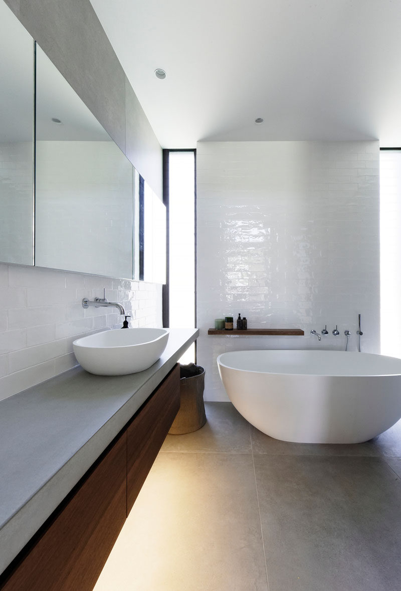 In this modern bathroom, a white freestanding bathtub has been placed beside a glossy tile wall, while a wood vanity and shelving adds a natural touch to the room. #ModernBathroom #BathroomDesign #Bathroom