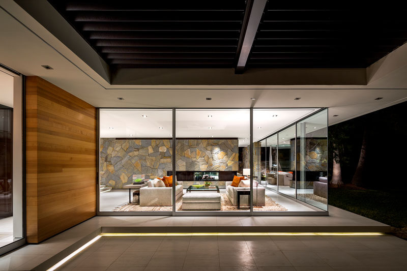 Floor to ceiling windows wrap around this modern living room, while a linear fireplace has been installed within the stone wall. #LivingRoom #StoneWall #ModernLivingRoom #GlassWalls