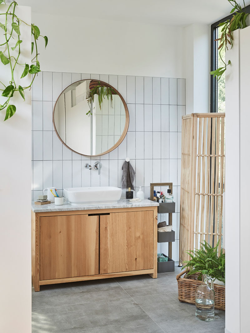 In this modern bathroom, plants add a touch of color to the space, that features white walls and tiles, a wood-framed round mirror, a wood vanity, and concrete flooring. #ModernBathroom #BathroomDesign