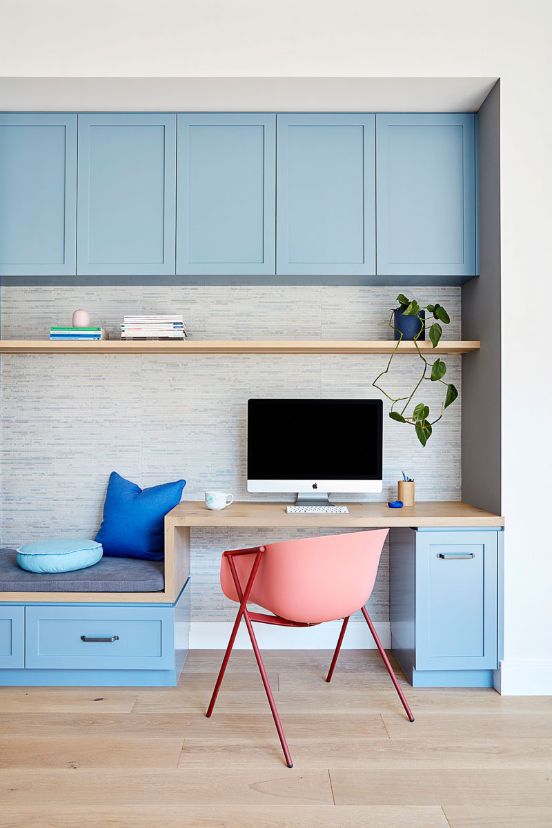 In this modern kitchen, matte light blue cabinets line the wall and transform into a small built-in home office with a bench, while a large white kitchen island provides additional seating. #LightBlueKitchen #ColorfulKitchen #KitchenIdeas #HomeOffice